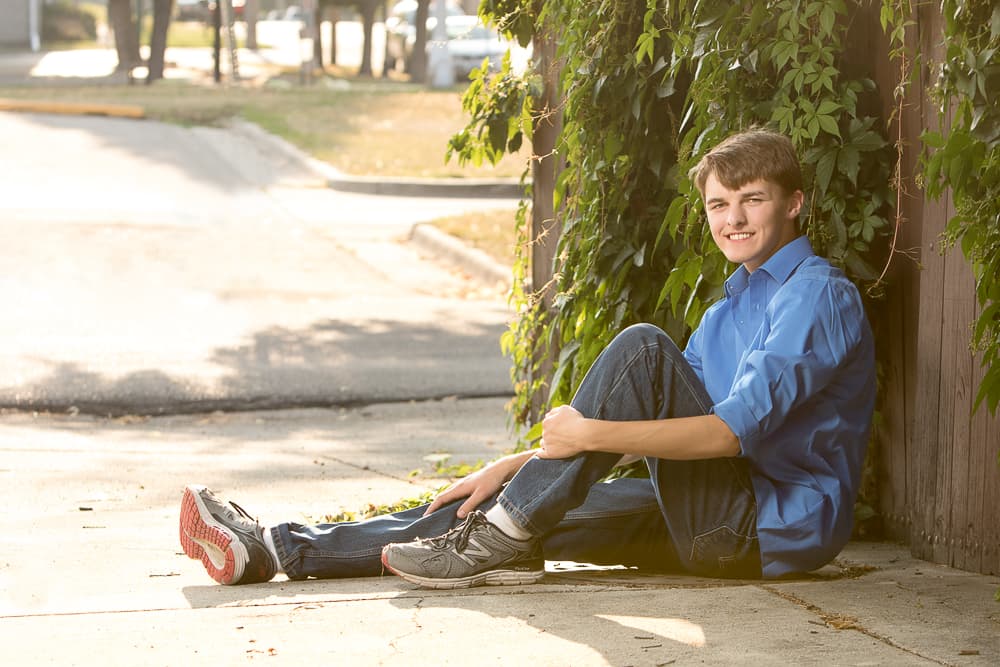 A senior sitting on a sidewalk against a wooden fence covered in ivy, backlit by late afternoon sun.