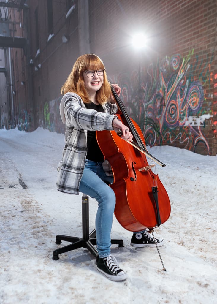 Ezra playing cello on a stool in a snow-covered alley, lit by a single strobe against a graffiti wall.
