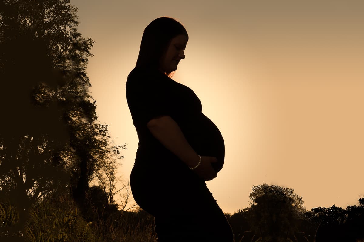 Jodine in profile silhouette against an amber sunset, hands under her pregnant belly.