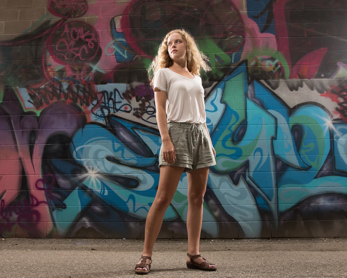 Oksana standing against a wall of blue and magenta graffiti, lit from the side in late daylight.