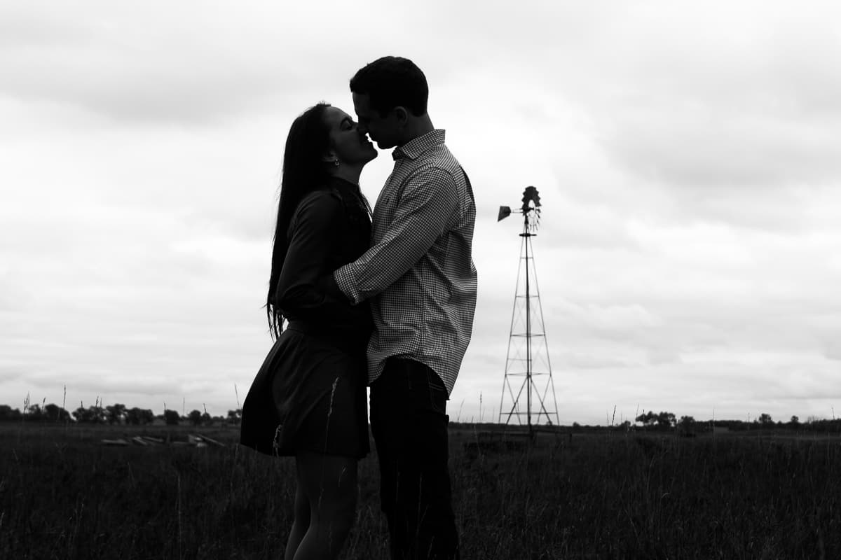 Stephanie and Eric nose to nose in a prairie field, silhouetted against a bright overcast sky with a windmill behind them.