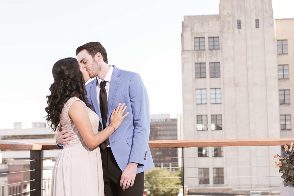 Karen and Taylor kissing on a downtown rooftop with an art deco tower behind them.