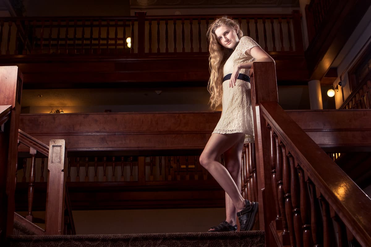 Tara leaning on the banister of a grand wooden staircase, lit warm against the dark hall behind her.