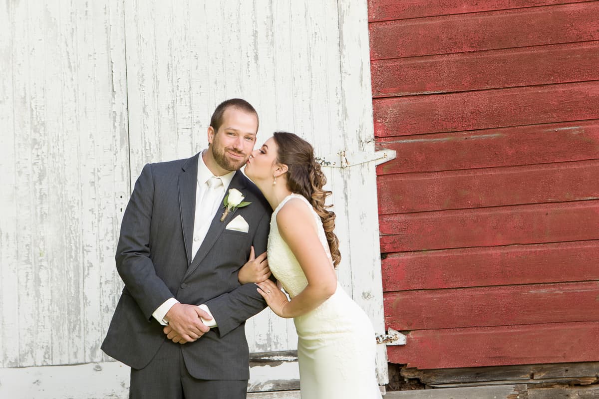 Tasha kissing Blake on the cheek in front of a weathered white and red barn door.