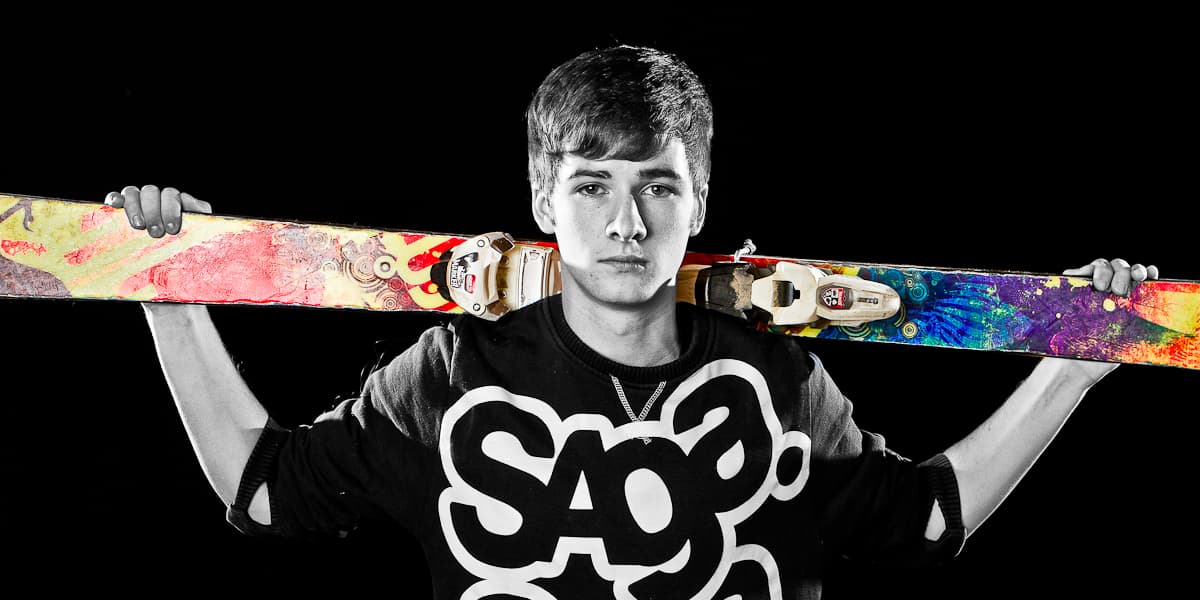 Ryan in black and white holding a pair of brightly painted skis across his shoulders, the skis left in full colour against a black background.
