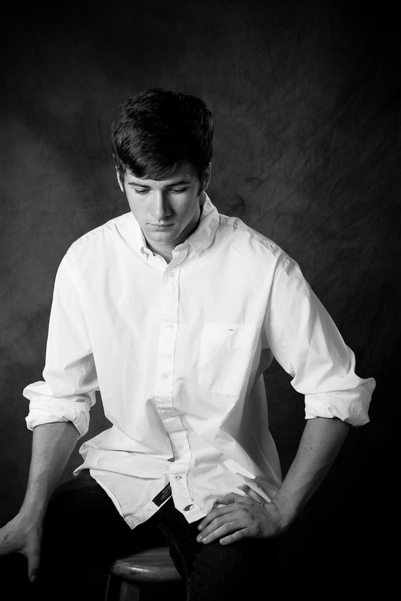 Ryan seated on a stool in a white shirt against a mottled backdrop, looking down, in black and white.