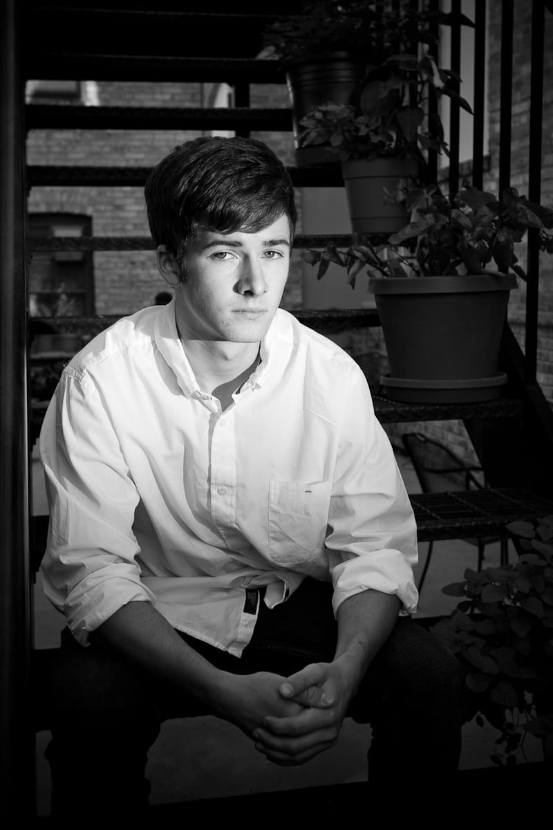 Ryan seated on iron steps beside potted plants and a brick wall, in black and white.