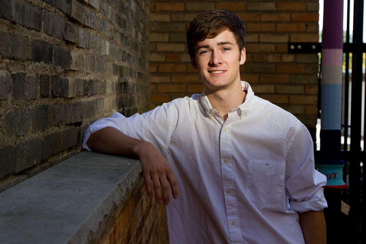 Ryan leaning on a concrete ledge against a brick wall in daylight, smiling at the camera.