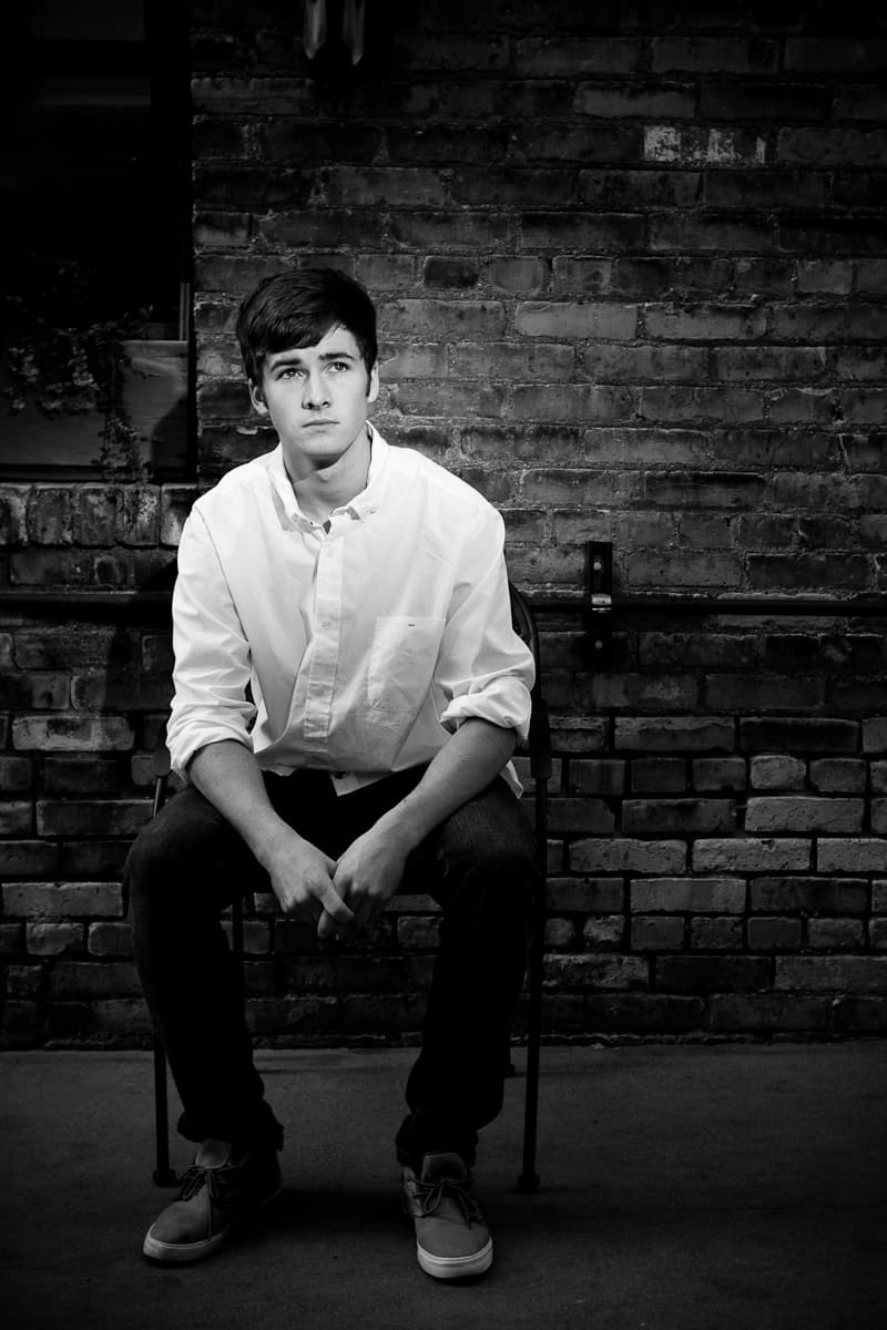 Ryan sitting on a folding chair against a weathered brick wall, hands clasped, in black and white.