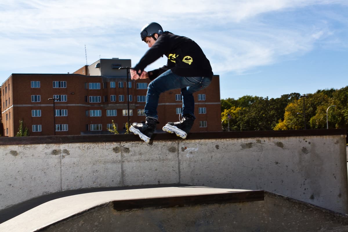 Ryan mid-air on inline skates above the lip of a concrete bowl, a brick building and blue sky behind him.