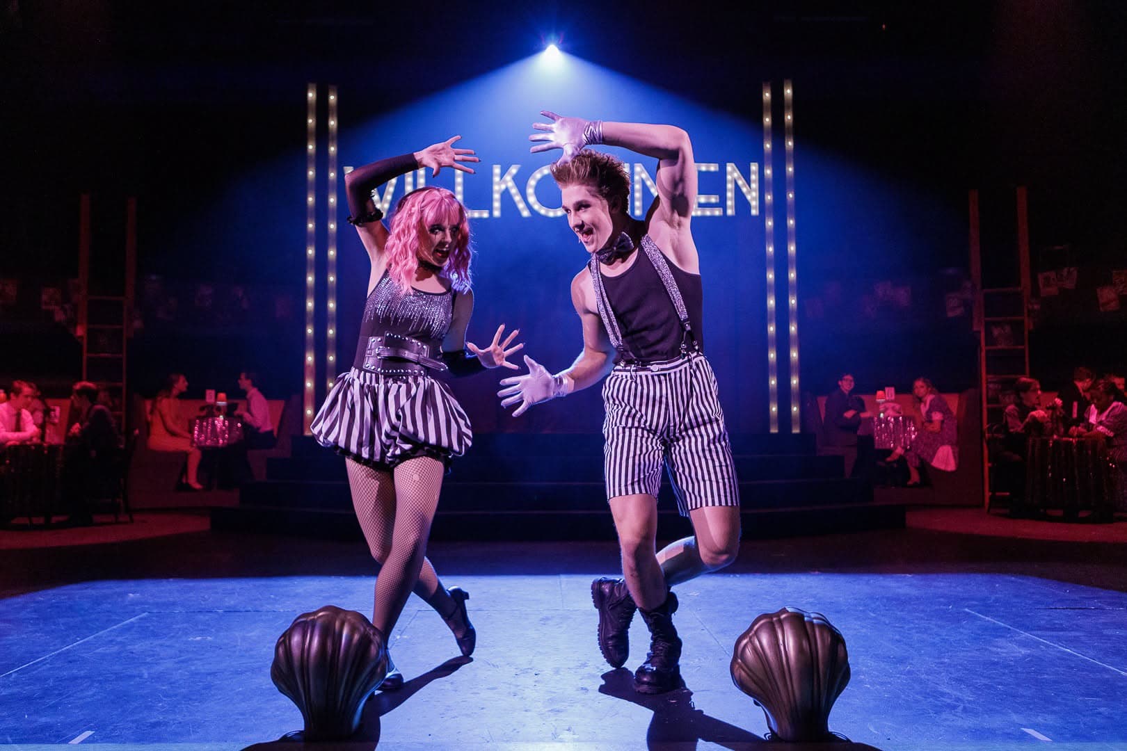 Two performers in striped costume framing their faces with gloved hands under a cool blue wash, the marquee dim behind them.