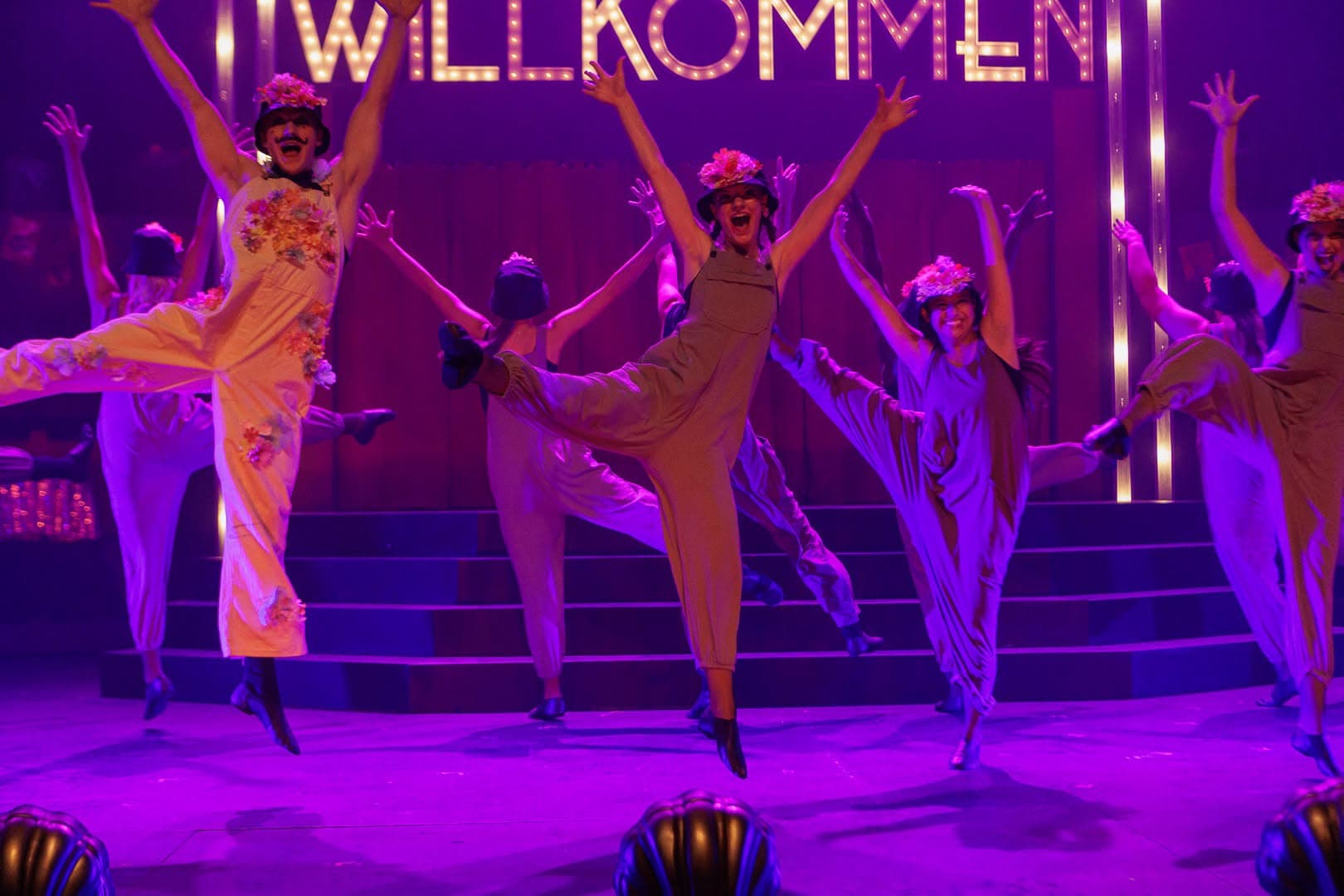 The whole company caught mid-jump in overalls and flowered hats, arms flung wide beneath the marquee.