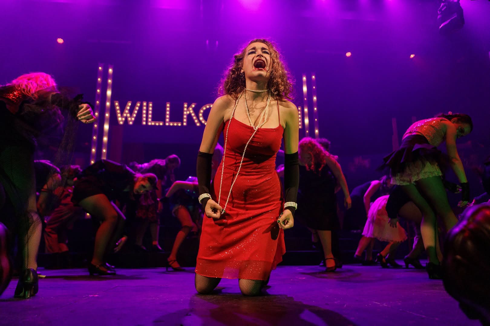 A performer in a red dress on her knees downstage, head back mid-note, the company bent low behind her in violet light.