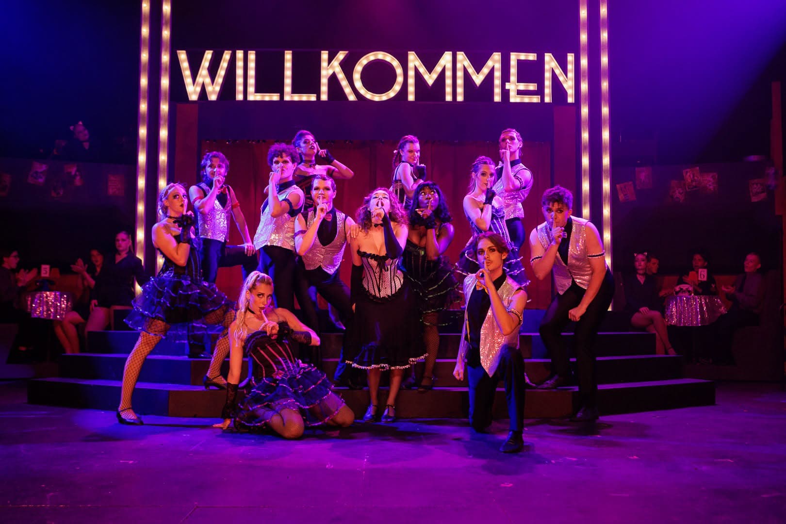 The full company posed on the stage steps with fingers to their lips, the lit WILLKOMMEN marquee spanning the wall behind them.