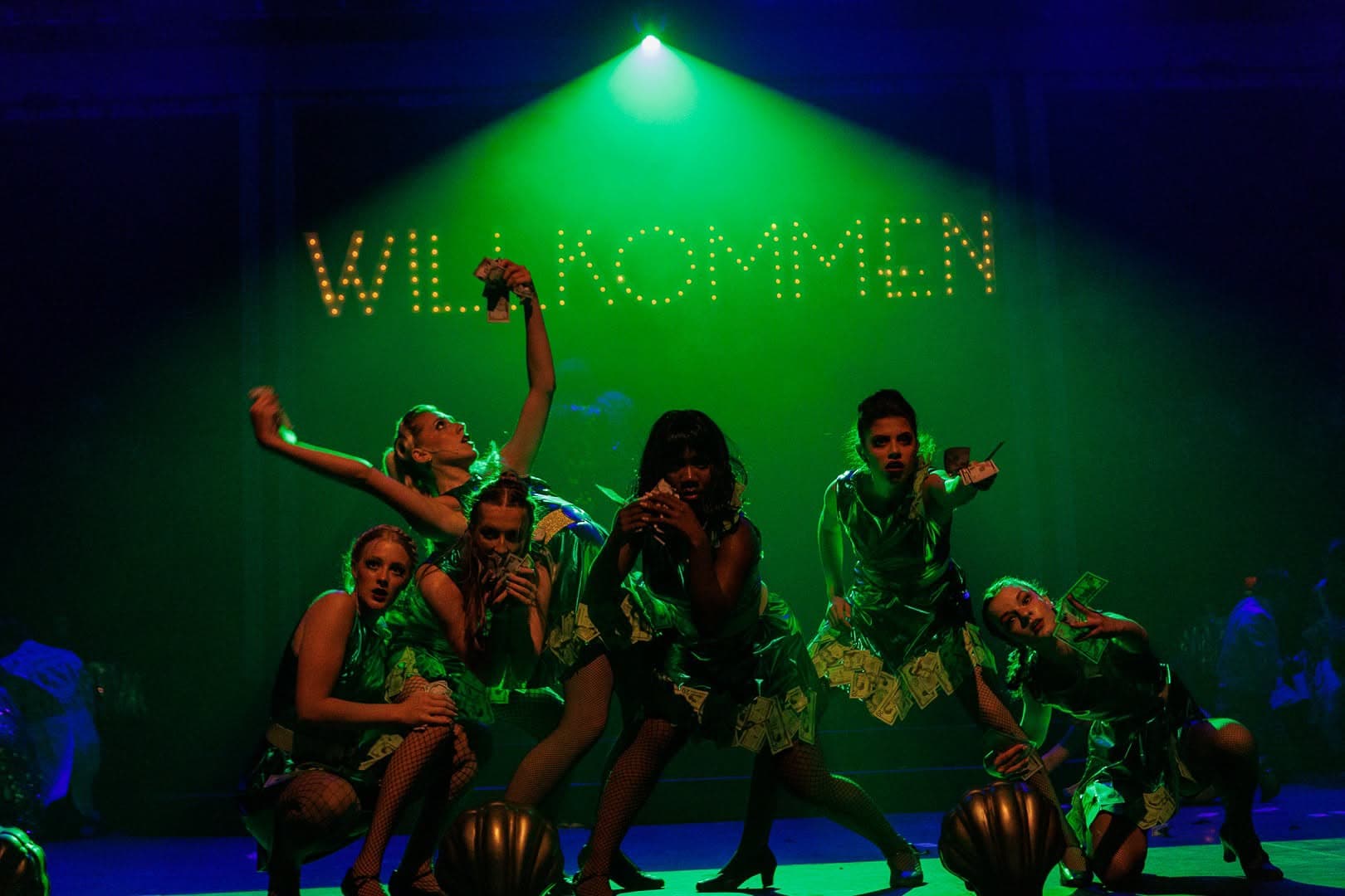 Six performers in green-lit costumes covered in banknotes, gathered under a single overhead beam with the WILLKOMMEN bulbs glowing behind them.
