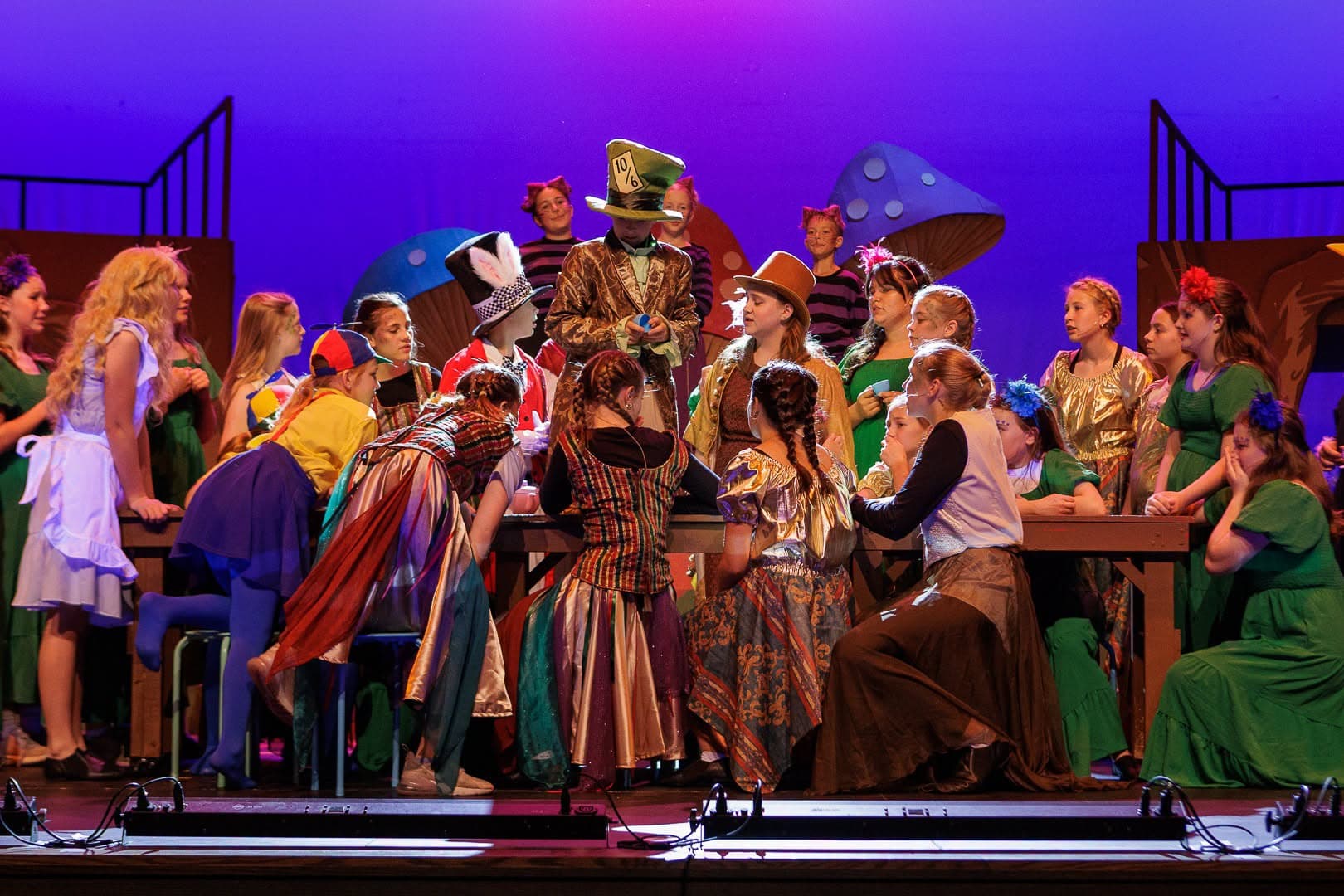 The full youth company crowded around the tea-party table under a purple wash, the Mad Hatter standing centre.