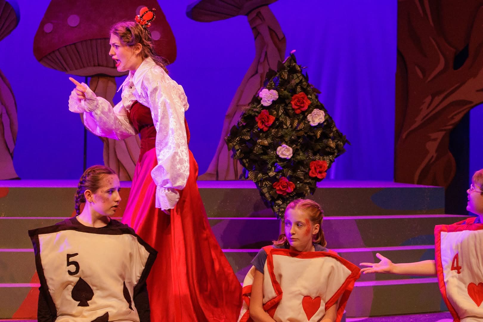 The Queen of Hearts in red mid-shout, pointing offstage, two playing-card guards seated below her.