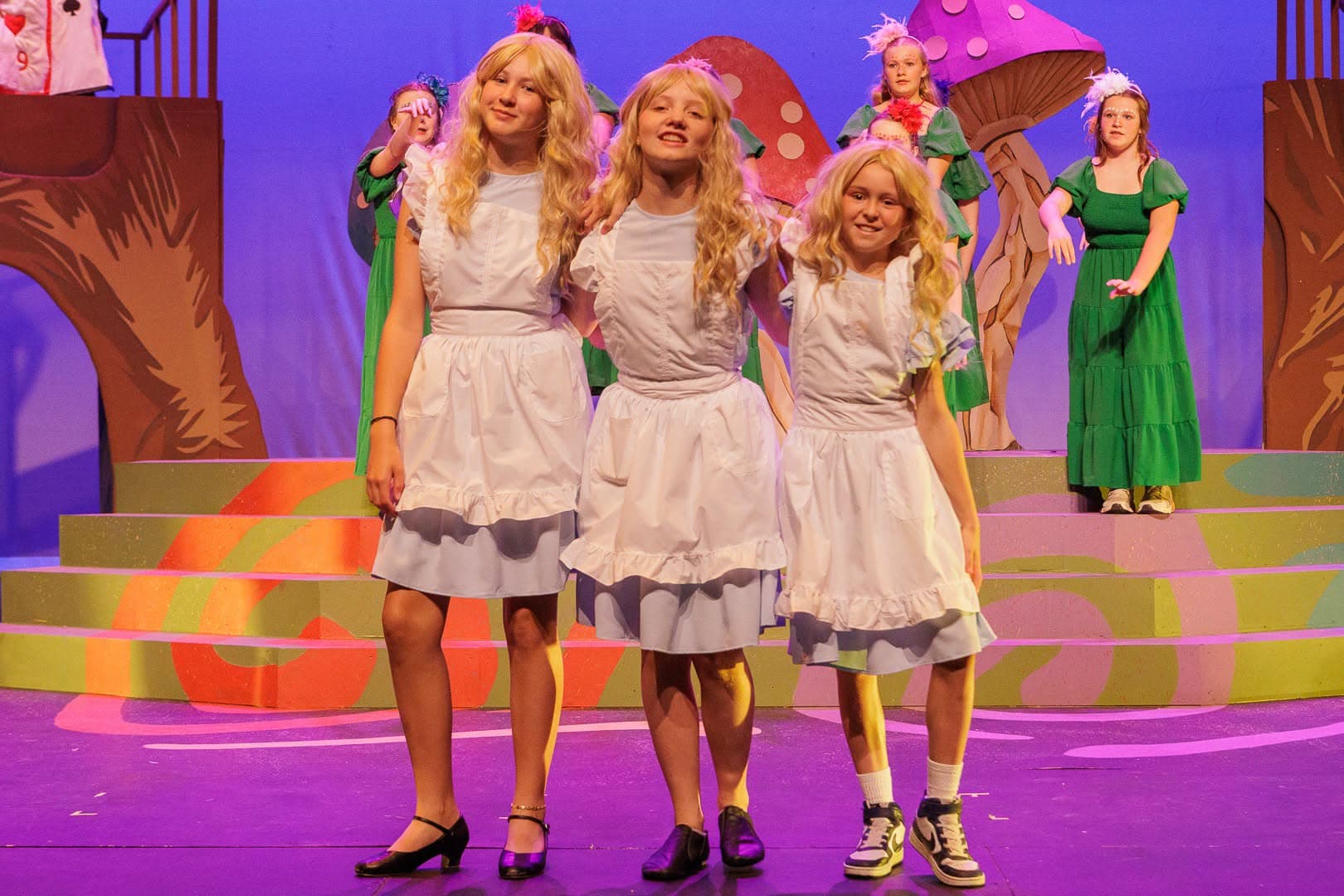 Three performers in matching Alice pinafores and blonde wigs standing shoulder to shoulder downstage.